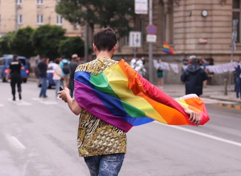person with short brown hair walking away down the street with rainbow flag around shoulders